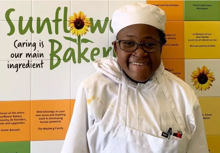 Student dressed in chef uniform poses in front of wall with Sunflower Bakery sign