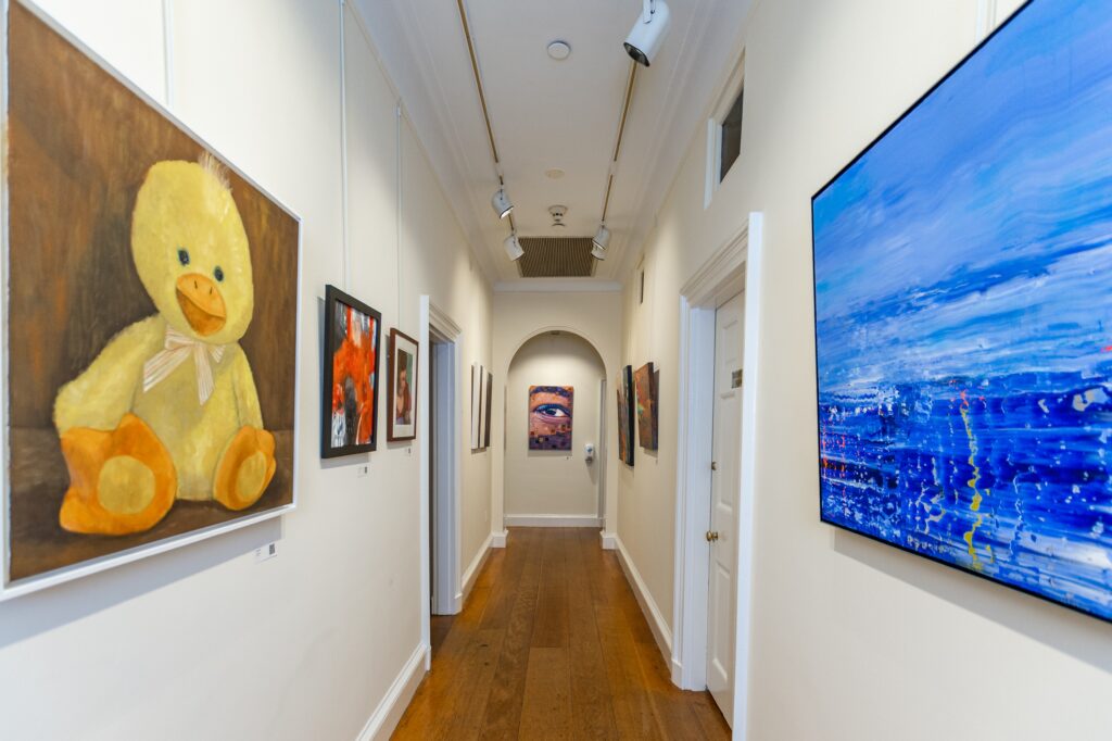 Art gallery corridor with painting of yellow, stuffed plush toy duck on the left and abstract blue and white painting on the right. The corridor ends with an arched doorway showing a painting of a human eye and eyebrow with abstract accents