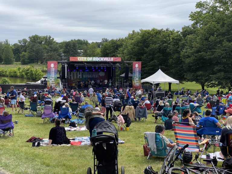 People seated outdoors in a park facing a stage with a banner in Rockville