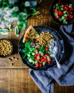 overhead photo of a bowl on a table containing chickpeas, vegetables, and a wedge of pita bread. a smaller bowl of salad is pictured in the upper right of the photo next to the larger bowl
